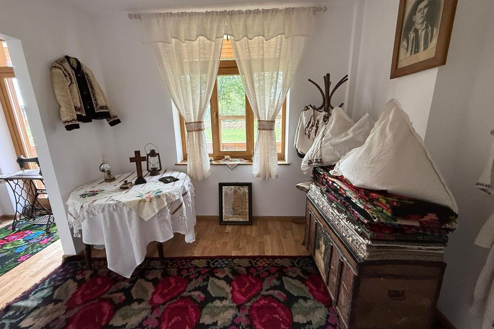 Sala de etnografie cu masa acoperită cu ștergar, patul cu lada de zestre și covorul cu trandafiri